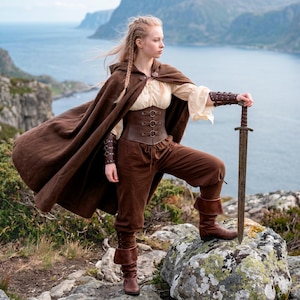 Könnte beinhalten: Eine Person in einem braunen mittelalterlichen Kostüm steht auf einem Felsen und hält ein Schwert. Das Outfit umfasst einen braunen Umhang, ein Korsett, eine Hose und Stiefel. Der Hintergrund zeigt eine malerische Aussicht auf das Meer und die Berge.