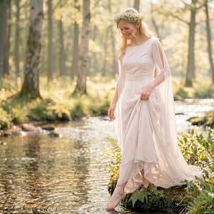 Puede incluir: Un vestido largo hasta el suelo de color blanco roto con mangas largas y fluidas abiertas y escote redondo. La prenda presenta un corpiño ajustado y una falda amplia con capas. Una delicada corona floral de pequeñas flores blancas.