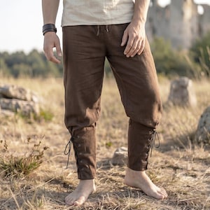 Op de afbeelding: Bruine linnen broek met een trekkoord in de taille en veterdetails bij de enkels. De broek is gemaakt van een zachte, getextureerde stof. Geschikt voor historische re-enactments of dagelijks gebruik. De stof is ademend.