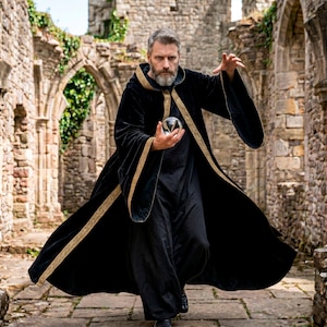 May include: A person in a black robe with gold trim, holding a crystal ball. The robe has wide sleeves and a hood. The background is a stone structure with archways and greenery.