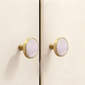 Puede incluir: Dos tiradores de armario con acabado dorado y centro redondo rosa claro. Los tiradores están montados en una puerta de armario color crema. Los tiradores tienen un diseño sencillo y elegante.