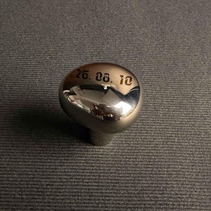 May include: A polished, rounded silver-toned metal knob featuring a smooth, reflective surface. The top of the knob is engraved with the black text 26. 08. 10 in a stylized font. The object has a rounded, ergonomic shape with a central base attachment.