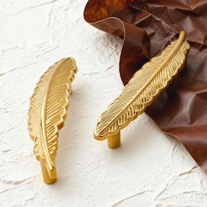 May include: Two gold-colored feather-shaped cabinet pulls. The pulls are made of metal and have a detailed feather design. The pulls are on a white surface with a brown paper background.