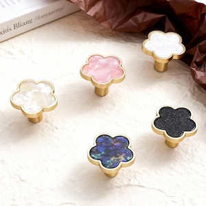 May include: Five flower-shaped cabinet knobs with gold-colored bases. The knobs feature various colors and textures, including white, pink, black, and blue. The knobs are arranged on a light-colored surface.