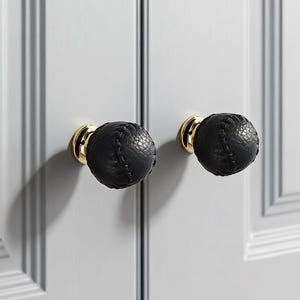 以下が含まれることがあります： 白いキャビネットドアに取り付けられた、金色のベースが付いた2つの黒い革製のキャビネットノブ。ノブは、ステッチのディテールが施されたテクスチャード加工の表面を備えており、キャビネットにエレガンスを添えています。