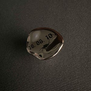 May include: A polished, irregular, silver-toned metallic object with a reflective surface. The surface features the black engraved text 26. 05. 10 in a gothic-style font. The object has a smooth, organic, rounded shape with subtle indentations.
