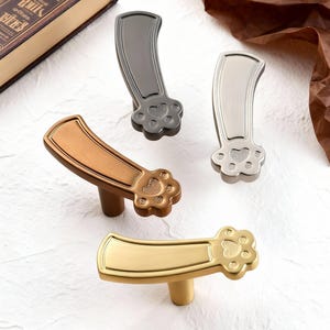 Könnte beinhalten: Vier Metall-Schrankgriffe in Bronze-, Gold-, Silber- und Grautönen. Jeder Griff hat ein Pfotenabdruck-Design an der Basis und einen gebogenen Griff. Die Griffe werden auf einer weißen Oberfläche präsentiert.