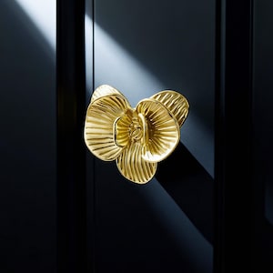 May include: A gold-coloured, flower-shaped cabinet pull against a black background. The pull has a detailed, textured surface, with the petals radiating outwards. The lighting creates shadows, highlighting the three-dimensional form of the pull.