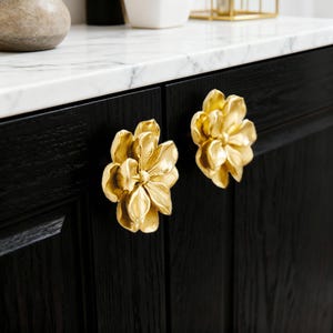 May include: Two gold flower-shaped cabinet knobs fixed to a black cabinet door. The knobs have detailed petal designs and a shiny finish. The cabinet door has a wood grain texture and is set against a white marble countertop.