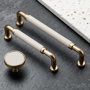 Op de afbeelding: Een set meubelbeslag met een gouden afwerking en crèmekleurige, getextureerde accenten. De set bevat twee stanggrepen en een ronde knop. Ontworpen voor meubels en kasten.