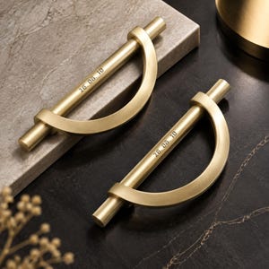 May include: Two brushed gold metal cabinet pulls, each featuring a straight horizontal bar connected to a semi-circular arch. The straight bar of each handle is engraved with the text 26. 06. 10 in a clear, sans-serif font style.
