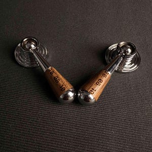 May include: Two matching furniture pulls featuring a polished chrome circular base and a tapered wooden handle. The wooden section of each pull is engraved with the date 26.06.10 in a dark, clear font, contrasting with the smooth, reflective metal.