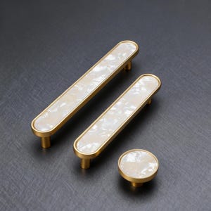 May include: A set of cabinet hardware featuring two long, rectangular pulls and a round knob. The hardware has a gold finish with a white, marbled inlay. The pulls are approximately 6 inches long, and the knob is about 1.5 inches in diameter.