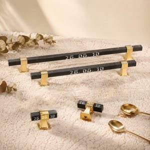 May include: Two long black cabinet pulls and two small black cabinet knobs, all featuring gold-toned metal accents and the engraved text 26. 05. 10. The hardware has a textured finish and is arranged on a soft, light-coloured surface.