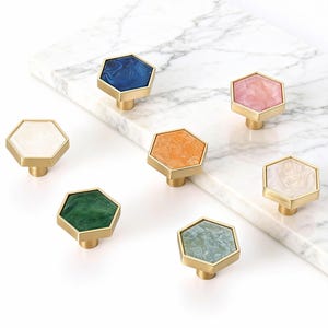 Könnte beinhalten: Sieben sechseckige Schrankknöpfe mit Messingbasen und bunten, marmorierten Oberflächen. Farben sind Blau, Rosa, Orange, Grün und Weiß. Die Knöpfe sind auf einer weißen Marmoroberfläche angeordnet.