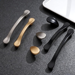 Könnte beinhalten: Eine Auswahl an Schrankgriffen und -knöpfen in Silber, Gold und Schwarz. Das Bild zeigt verschiedene Stile von Griffen und Knöpfen mit einem modernen Design. Die Hardware wird auf einer dunklen, strukturierten Oberfläche präsentiert. Im Hintergrund ist ein weißes Tablett zu sehen.