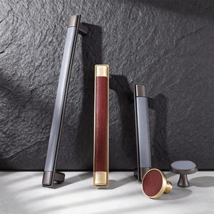 Pode incluir: Uma coleção de puxadores de armário e um botão. Os puxadores são cinza e preto, com um puxador de couro vermelho e dourado. O botão tem uma face de couro vermelho. Apresentado em um fundo escuro e texturizado.