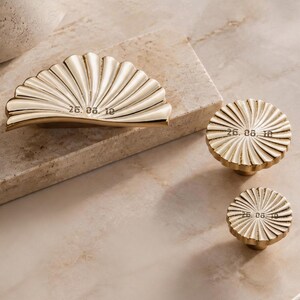 May include: A set of three gold-toned metal hardware pieces featuring a fan-shaped handle and two circular knobs. Each piece has a ribbed, sunburst-like texture and is engraved with the date 26. 08. 10 in a dark, stylised font on the surface.