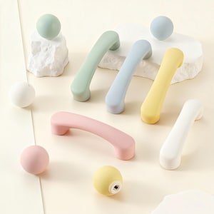 Può includere: Una collezione di maniglie e pomelli per armadi colorati in tonalità pastello di verde, blu, giallo, rosa e bianco. Le maniglie sono curve e i pomelli sono rotondi. L'hardware è disposto su una superficie chiara.