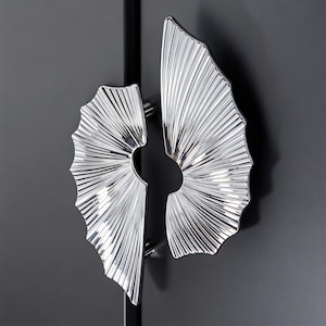 May include: Close-up of two silver cabinet handles with a fan-like design. The handles are mounted on a dark grey cabinet door. The handles have a textured, ridged surface, and a circular opening in the centre.