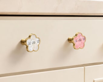 May include: Two flower-shaped drawer knobs with a gold-toned metal base. One knob features a white pearlescent inlay, and the other has a pink pearlescent inlay. Both knobs are engraved with the black text 26. 08. 10 on their front surfaces.