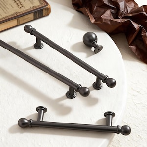 Könnte beinhalten: Eine Sammlung von dunkler Bronze-Schrankbeschlägen, darunter Stangengriffe und ein Knauf. Die Griffe haben ein schlichtes, elegantes Design mit abgerundeten Enden und dekorativen Basen. Die Hardware wird auf einer weißen Oberfläche präsentiert.