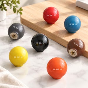 May include: A collection of seven spherical cabinet knobs in red, blue, gray, black, yellow, brown, and orange. Each knob features a smooth, glossy finish and is printed with the white text 26. 08. 18. The knobs include a central metal screw insert.