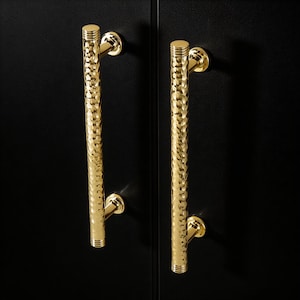 Puede incluir: Dos tiradores dorados para armarios con un acabado texturizado y martillado. Los tiradores son cilíndricos con una parte superior e inferior redondeadas, montados sobre una superficie negra. El herraje tiene un diseño moderno y elegante. Los tiradores miden unos 20 cm de largo.