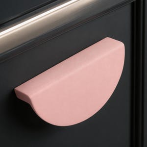 Può includere: Una maniglia per armadio rosa chiaro, a forma di semicerchio. La maniglia è montata su un'anta di armadio grigio scuro. Una finitura metallica color argento è visibile sopra la maniglia. Il design della maniglia è moderno e minimalista.