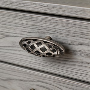 Op de afbeelding: Close-up van een grijze houten lade met een ovale metalen handgreep. De handgreep heeft een roosterontwerp en een zilveren afwerking. De houtnerf is zichtbaar en voegt textuur toe aan het stuk. De handgreep is ongeveer 7 cm lang.