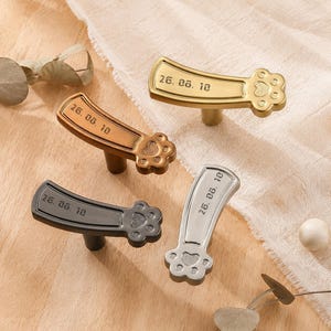 May include: Four metal drawer knobs in gold, bronze, black, and silver finishes. Each knob features a curved handle shape with a paw print design at one end and the engraved text 26. 08. 10 on the surface. The knobs have a central post.