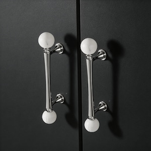 May include: Two cabinet pulls with a polished chrome finish and white marble sphere accents. The pulls are mounted on a dark gray cabinet door. The design features a long, slender handle with a decorative sphere at each end.