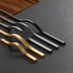 May include: A collection of modern cabinet pulls in various finishes. The pulls have a wavy, curved design with a rectangular profile. Finishes include gold, dark gray, and black, arranged diagonally against a dark background.