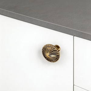 Peut inclure: Gros plan sur un bouton de meuble doré avec un motif en forme d'éventail. Le bouton est fixé à une porte d'armoire blanche, avec un plan de travail gris en arrière-plan. Le design du bouton présente des lignes rayonnantes.