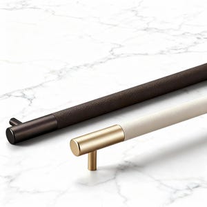 Puede incluir: Dos tiradores de armario. Uno es marrón oscuro con un extremo negro, el otro es blanquecino con un extremo dorado. Ambos tienen un diseño cilíndrico y están sobre un fondo de mármol blanco. Para muebles o armarios.
