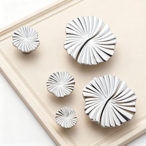 May include: A collection of silver-colored cabinet knobs and pulls. The knobs are round with a radial design, while the pulls have a stylized petal shape. The hardware is displayed on a cream-colored surface.