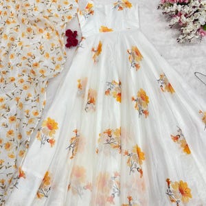 Puede incluir: Vestido blanco con tirantes finos y falda fluida, estampado con flores amarillas y naranjas. Un chal floral a juego es visible a la izquierda. El vestido probablemente está hecho de una tela ligera y transparente.
