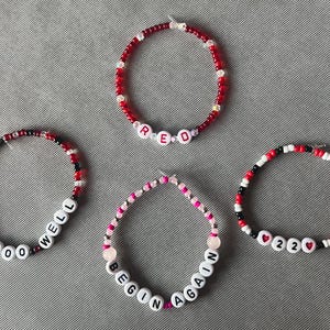 May include: Four circular beaded bracelets featuring small red, black, white, and pink beads. One bracelet spells RED, another ALL TOO WELL, one says BEGIN AGAIN, and the last features 22 with heart icons. The beads are arranged in various patterns.