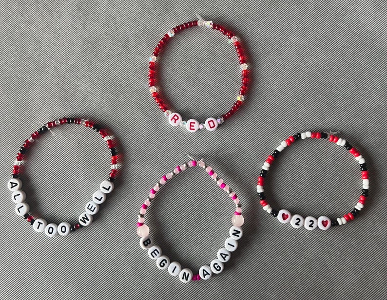 May include: Four circular beaded bracelets featuring small red, black, white, and pink beads. One bracelet spells RED, another ALL TOO WELL, one says BEGIN AGAIN, and the last features 22 with heart icons. The beads are arranged in various patterns.