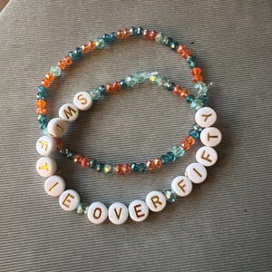 May include: Two beaded bracelets featuring faceted orange, teal, and light blue glass beads. One bracelet includes white round beads with gold lettering that spells out the phrase I'M FIFTY OVER FIFTY, arranged in a circular pattern.