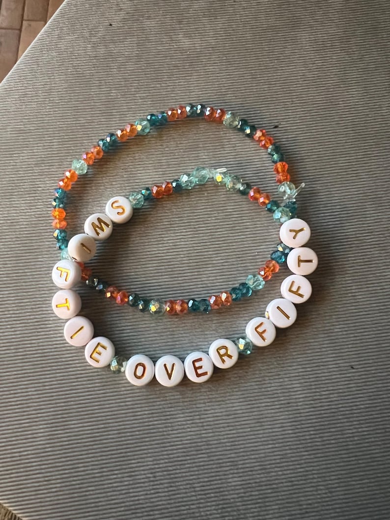 May include: Two beaded bracelets featuring faceted orange, teal, and light blue glass beads. One bracelet includes white round beads with gold lettering that spells out the phrase I'M FIFTY OVER FIFTY, arranged in a circular pattern.