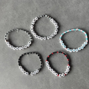 May include: Five circular beaded bracelets featuring white letter beads spelling out THE TORTURED POETS DEPT, FORTNIGHT, THE PROPHECY, DOWN BAD, and GUILTY AS SIN. The bracelets are constructed with various colored plastic beads including black, red, and pastel tones.