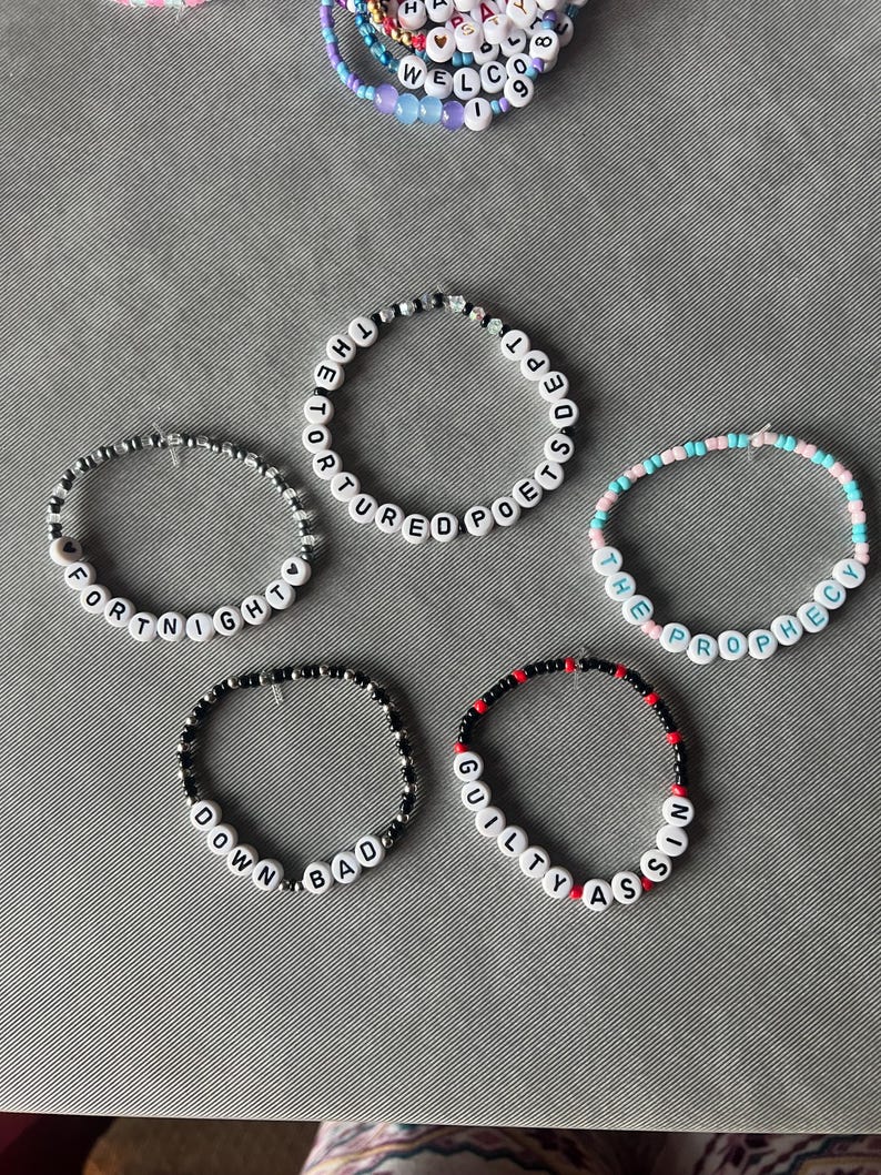 May include: Five circular beaded bracelets featuring white letter beads spelling out THE TORTURED POETS DEPT, FORTNIGHT, THE PROPHECY, DOWN BAD, and GUILTY AS SIN. The bracelets are constructed with various colored plastic beads including black, red, and pastel tones.