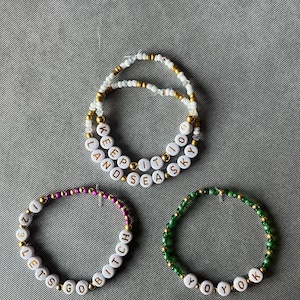 May include: Three beaded bracelets featuring white letter beads and metallic gold accents. One bracelet displays the text KEEP IT TO LAND SEA SKY, another shows 1 2 3 LETS GO BITCH, and the third features YOYOK. A partial bracelet with WELCOME is visible.