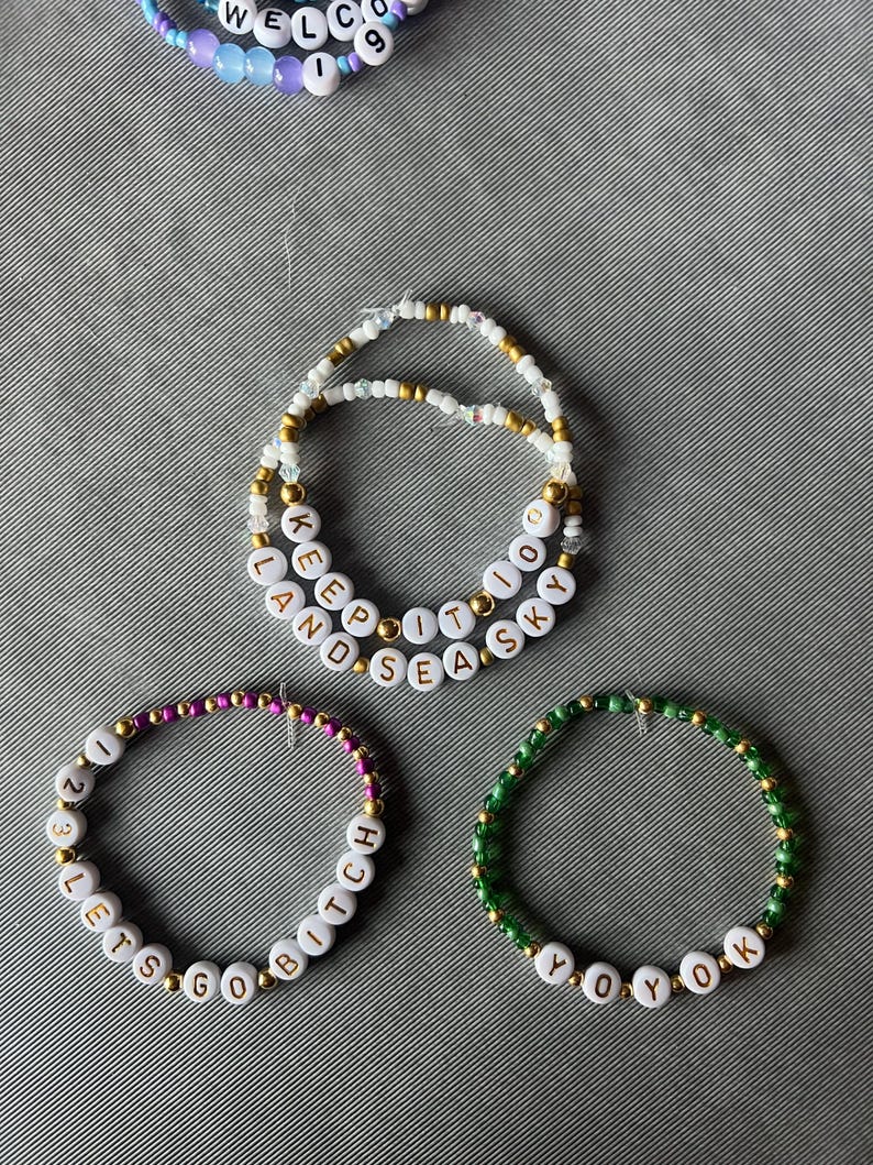 May include: Three beaded bracelets featuring white letter beads and metallic gold accents. One bracelet displays the text KEEP IT TO LAND SEA SKY, another shows 1 2 3 LETS GO BITCH, and the third features YOYOK. A partial bracelet with WELCOME is visible.