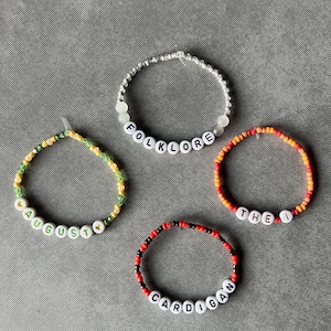 May include: Four circular beaded bracelets featuring white letter beads. One bracelet spells FOLKLORE in silver and white beads, one spells AUGUST with yellow heart beads in green and yellow, one spells THE I in orange and red, and one spells CARDIGAN in red and black.