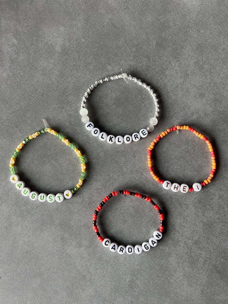 May include: Four circular beaded bracelets featuring white letter beads. One bracelet spells FOLKLORE in silver and white beads, one spells AUGUST with yellow heart beads in green and yellow, one spells THE I in orange and red, and one spells CARDIGAN in red and black.