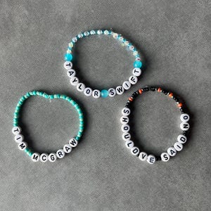May include: Three beaded bracelets featuring white letter beads and colorful accents. One bracelet displays the text Taylor Swift, another reads Tim McGraw, and the third features the text Should've Said No, all arranged in circular patterns.