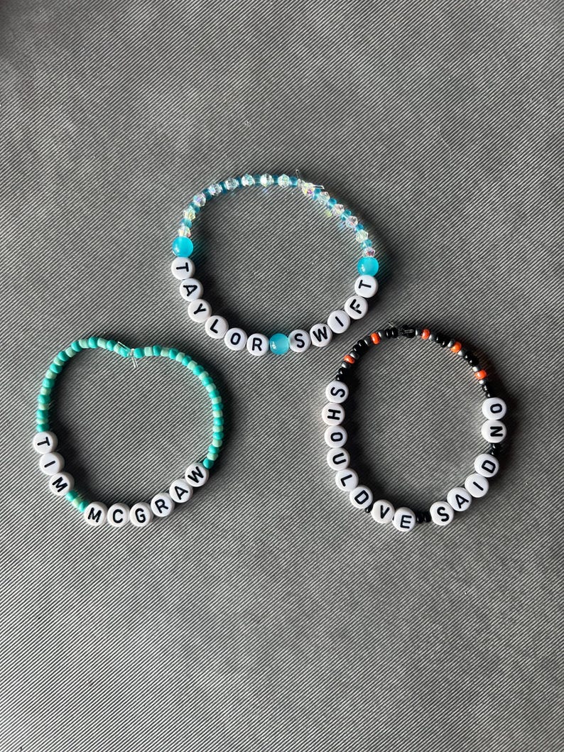 May include: Three beaded bracelets featuring white letter beads and colorful accents. One bracelet displays the text Taylor Swift, another reads Tim McGraw, and the third features the text Should've Said No, all arranged in circular patterns.