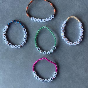 May include: Five beaded bracelets featuring white letter beads spelling out EVERMORE, TIS THE DAMN SEASON, WILLOW, CHAMPAGNE PROBLEMS, and TOLERATE IT. Each bracelet uses different colored seed beads including brown, blue, green, cream, and pink.
